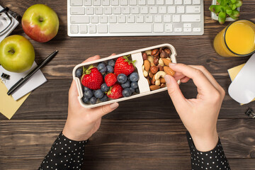 First person top view photo of woman's hands holding lunchbox with healthy meal nuts and berries...