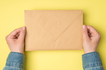 First person top view photo of female hands holding closed craft paper envelope on isolated yellow...