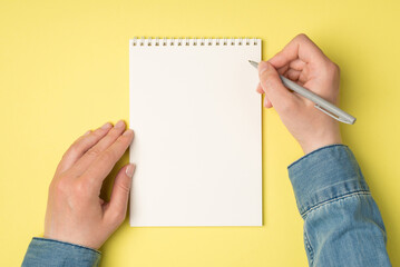 Obraz premium First person top view photo of female hands holding pen over open planner on isolated yellow background with blank space
