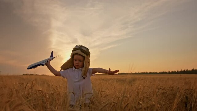 Slow-motion, happy boy in hat and aviation pilot glasses playing with an airplane in his hands at sunset run on meadow wheat field. Child dreams becoming aviation pilot, plays with toy airplane.