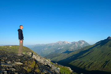 A man by profil, looking green mountains. A hiker contemplates the pastures and snow-capped peaks in summer at dawn.
