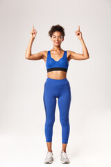 Fototapeta na wymiar Full length of smiling attractive Black sportswoman in blue sports clothing, looking pleased at camera and pointing fingers up, showing logo or advertisement about workout, white background