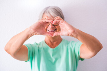 Portrait of one mature and old woman showing to them camera a heart made with her hands smiling and having fun at home. Female senior taking care and loving people
