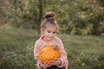 Happy smilling child girl in a knitted sweater is holding in hands ugly orange pumpkin. Deformed...