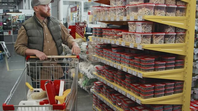 Slowmo handheld shot of bearded man in puffy vest and cap picking up home improvement supplies and putting it into shopping cart at hardware store