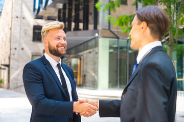 Confident businessman and his colleague are shaking hands in front of modern office building. Financial investors outdoor. Banking and business.
