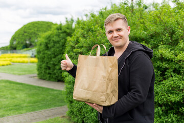 Man with eco paper bag for takeaway food showing thumbs up on nature green background. Delivery in...