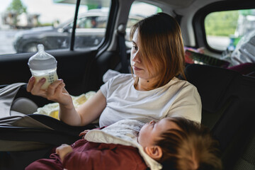 Ydult caucasian woman female mother sitting on the back seat of her car holding formula or breast milk in baby bottle ready to feed her child while traveling on the road in summer day on holiday
