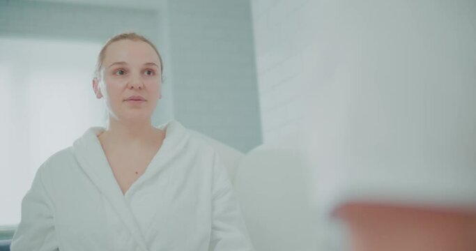A close-up female client is sitting in the cosmetology clinic in a bathrobe and looking at somebody in front of her.