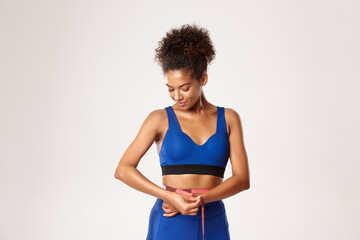 Fototapeta na wymiar Smiling african-american female athlete in blue leggings and sportbra, measuring her waist and hips with tape, standing over white background