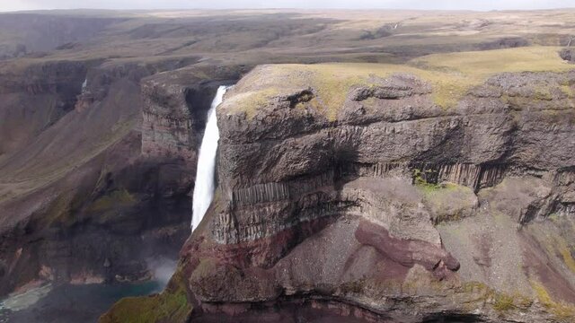 Aerial shot moves around cliff to reveal tall waterfall behind. Cloud shadows move creating dramatic landscape. Shot in Iceland.