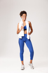 Full length of happy and healthy african-american fitness woman, wearing blue outfit, standing with water bottle and towel after exercises, posing over white background