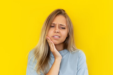 Close-up of a young caucasian beautiful blonde woman with a toothache holding her cheek isolated on color yellow background. The girl suffers from a toothache