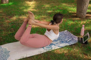 Vertical image of a young woman doing the bow pose while making a video with her tablet for her social media. Concept of outdoor pilates.