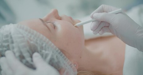 Close-up female client in disposable hat and cosmetologist is using a cosmetic pencil for painting white stripes on her cheek.