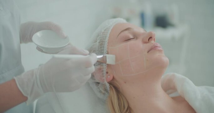 The female client is laying wearing a disposable hat and on her face are painted with white squares, cosmetologist is applying a cream onto her face by using a cosmetic brush.