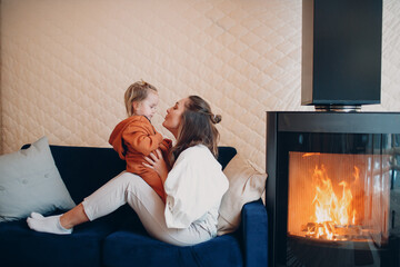 Mother and child sitting and playing on sofa near fireplace. Single parenting. Mom and baby child. Parent and little kid relaxing at home.