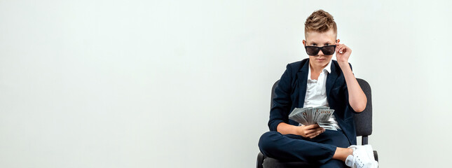 A rich boy on a light background holds a bundle of dollars in his hands. Financial education, young millionaire, investments, loans, winnings, dividends. Flyer blank, site header, copy space.