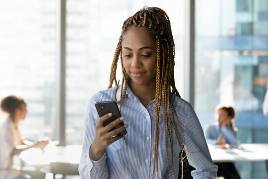 Happy beautiful young african ethnicity multiracial businesswoman manager employee looking at smartphone screen, reading message with good news, web surfing information, using software applications.