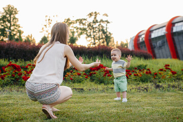 A young cute mother helps and teaches her little son to take his first steps during sunset in the...