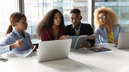 Fototapeta premium Group of concentrated young african american business people coworkers colleagues looking in paper planner, discussing ideas, analyzing sales statistics, sitting together at table in modern workplace.