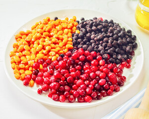Frozen berries. Immune boosting concept. Organic, frozen sea buckthorn, cranberry and blueberry berries on a white plate and a jar of honey on a white background. Cooking homemade berries mors or jam © KRISTINA KUPTSEVICH