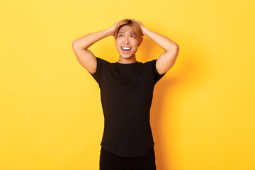 Portrait of young asian guy panicking, looking anxious and upset, holding hands on head and grimacing, standing over yellow background