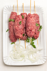 Raw minced meat on wooden skewers and chopped onion rings on white plate.