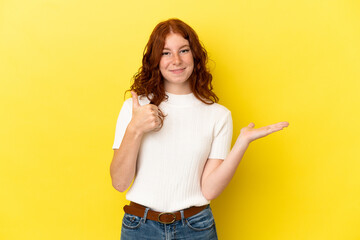 Teenager reddish woman isolated on yellow background holding copyspace imaginary on the palm to insert an ad and with thumbs up