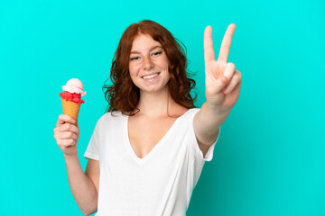 Teenager reddish woman with a cornet ice cream isolated on blue background smiling and showing...