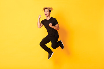 Fototapeta na wymiar Full-length of happy attractive asian guy in black clothes jumping and celebrating victory, achieve goal, standing yellow background, triumphing