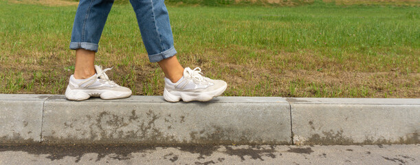 A woman in jeans and sneakers walks along the curb. Legs in sneakers close-up, banner.