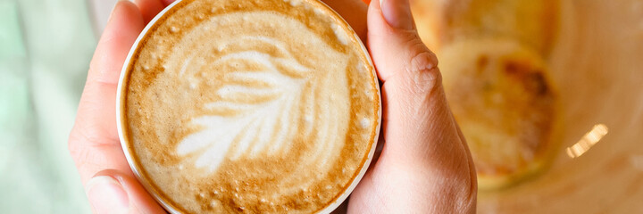 female hands holding a cup of fresh brewed coffee with beautiful leaf latte art from foam on the background of the table with a plate of food. the concept of morning breakfast and brunch. banner
