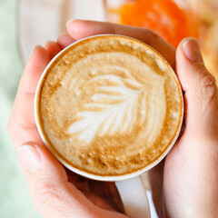 female hands holding a cup of fresh brewed coffee with beautiful leaf latte art from foam on the background of the table with a plate of food. the concept of morning breakfast and brunch. square
