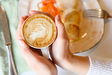 female hands holding a cup of fresh brewed coffee with beautiful leaf latte art from foam on the background of the table with a plate of food. the concept of morning breakfast and brunch