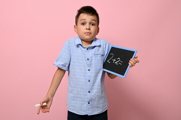 A cute schoolboy holds a blackboard in his hand with an arithmetic problem and with surprise and...