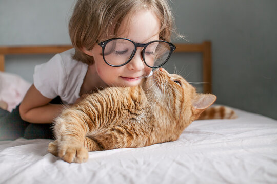 little girl with glasses lies on the bed and hugs a fat ginger cat. High quality photo