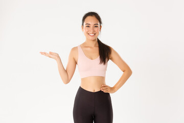 Sport, wellbeing and active lifestyle concept. Portrait of cheerful attractive asian fitness coach, female athlete introduce or demonstrate product for workout, holding something on hand
