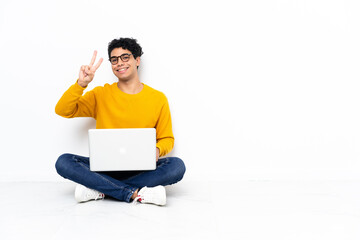 Venezuelan man sitting on the floor with laptop smiling and showing victory sign
