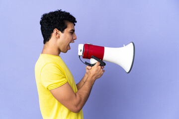 Young Venezuelan man isolated on purple background shouting through a megaphone to announce something in lateral position