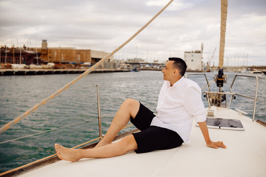 A man sits on the bow of a sailing yacht and enjoys life. Handsome guy in a white shirt, businessman, freelancer resting in the sea or ocean