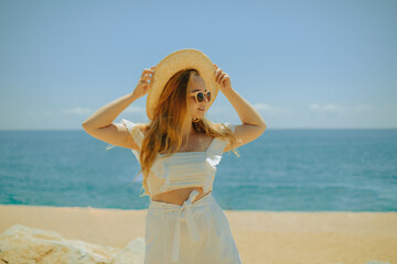 Fototapeta na wymiar Beautiful girl on the background of the beach, sea and ocean. A blonde in a white outfit on vacation, wearing sunglasses and a larger hat, enjoying a vacation in the summer.