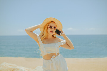 Fototapeta na wymiar A beautiful girl on the background of the beach, sea and ocean is calling. A blonde in a white outfit on vacation speaks on the phone, roaming services, freelancer, earning money on vacation, summer.