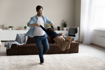 Happy father and excited little son enjoying funny active energetic games in living room, playing swirl and sway. Dad spinning around, holding arms of kid flying in air, having fun at home.