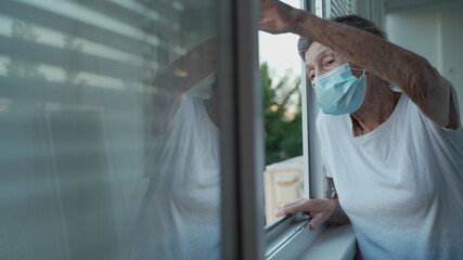 Happy senior woman in mask waves to family from window hospital. Masked elderly female glad that children came visit her in nursing home. Grandmother greets child with hand of relatives at clinic