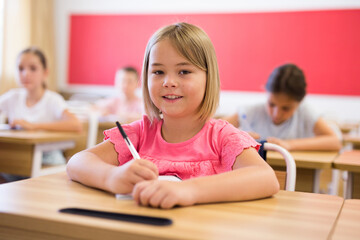 Portrait of cheerful blonde tween girl studying in classroom, listening to schoolteacher and writing in notebook