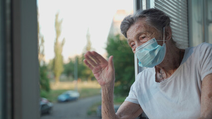 Portrait of masked senior woman in late 90s waving from home window. Elderly woman in protective mask looks out the window and waves her hand while isolating at home and conducting social distancing