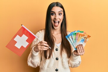 Beautiful brunette young woman holding switzerland flag and franc banknotes celebrating crazy and amazed for success with open eyes screaming excited.