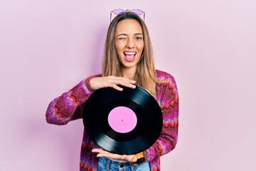 Beautiful hispanic woman wearing hippie style holding vinyl disc winking looking at the camera with...