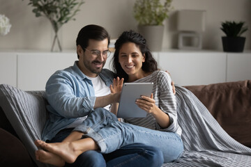 Happy joyful 30s couple resting on couch at home, hugging, using virtual order app on tablet computer, making video call, payments, shopping and buying online, browsing internet, watching content